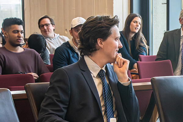 A lawyer sits in a courtroom and places his hand on his chin.