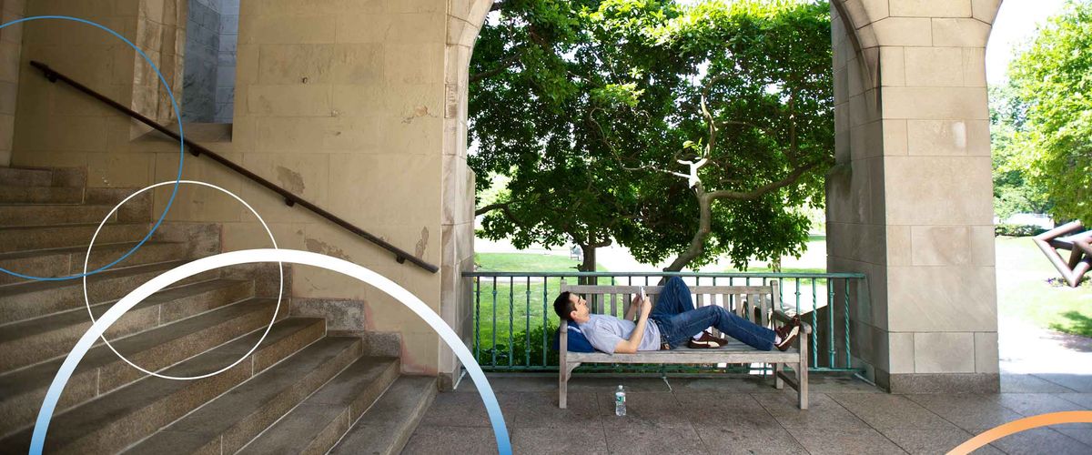 A Boston University student reclining and reading on a bench outdoors