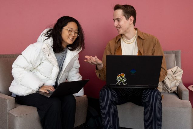 Photo: A young Asian woman wearing glasses and a white puffer jacket leans to the right to look at the screen of a young white man's laptop which is sitting in his lap. He looks over to his right at the woman and smiles and gestures knowingly. They both sit in front of a pinkish wall.