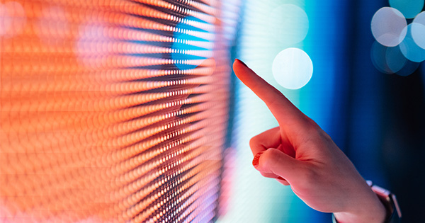 An outstretched finger pointing towards a digital screen lit up with oranges, reds, and blues