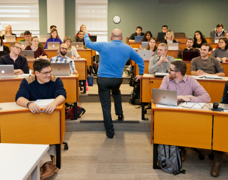 Professor in classroom, standing between rows of seated students, raising his hand for emphasis