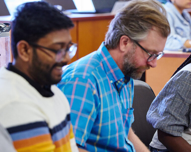 Four students sitting in a row in a classroom with the only female student leaning forward and engaging with the three male students.