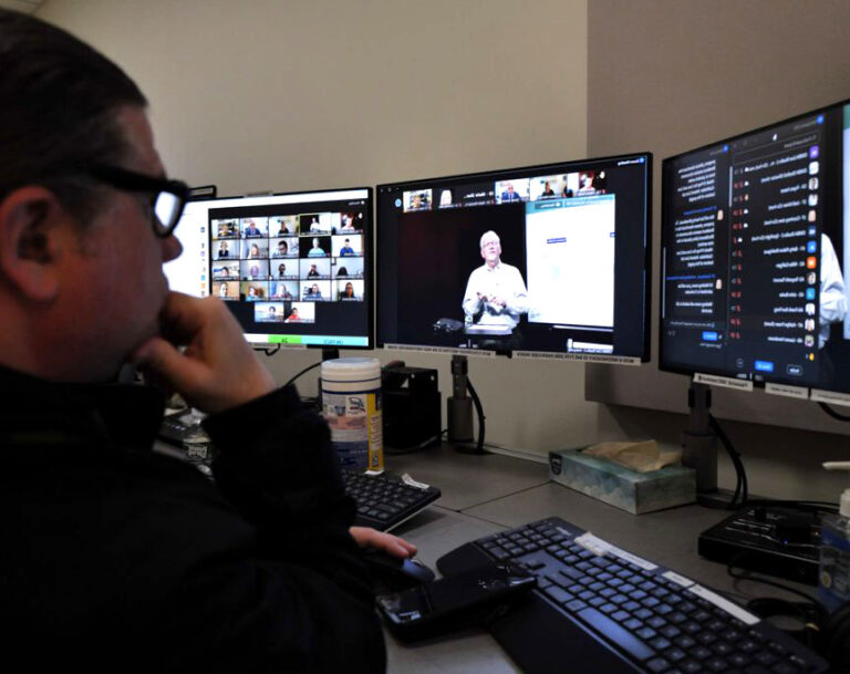 Person sitting in front of multiple screens managing the technology while a professor teaches an online class in a studio.