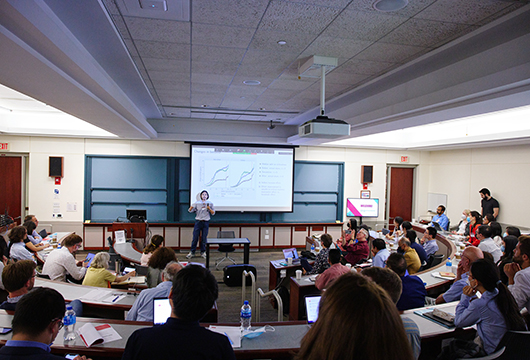 Professor standing in front of a large classroom of students with a projector displaying a slide behind her
