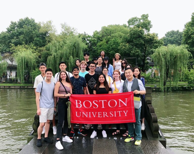 Group of over 10 study abroad students hold a BU sign and pose on a dock overlooking a small body of water with lush draping trees.