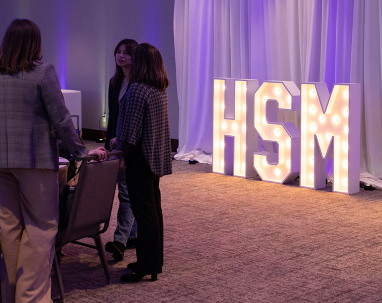 Students stand in a darkened conference room with HSM 50 in large light up letters after a Health Sector event.