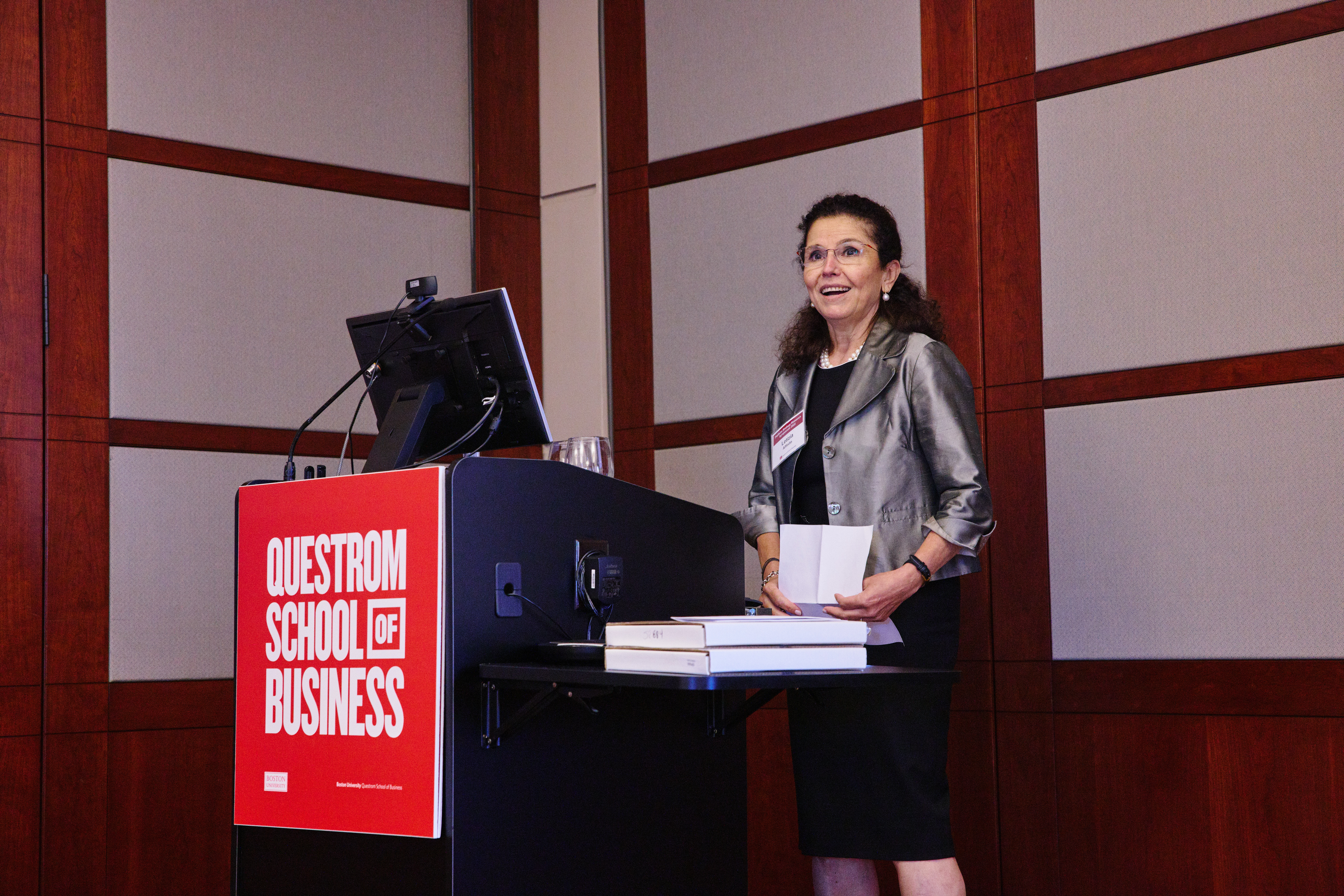 Woman with shiny grey jacket and brown curly hair smiling and presenting at the podium which has a Questrom Means Business logo on it