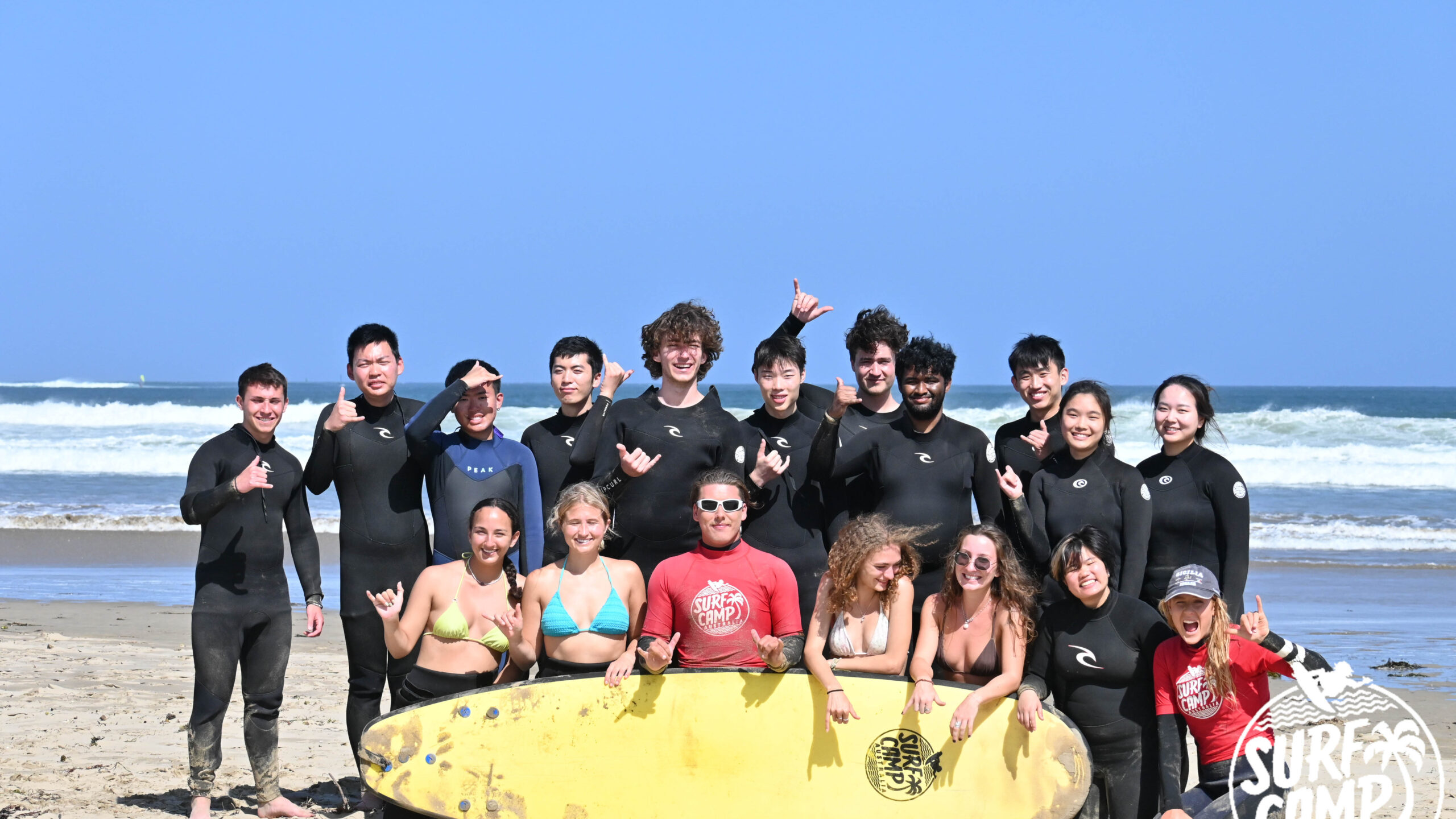 A group of over 10 study abroad students stand together on the beach with surfing attire on.