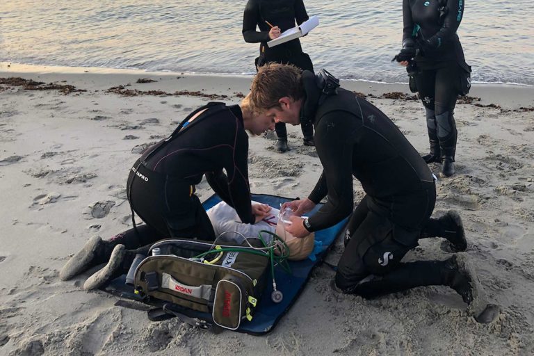 Two people on a beach in wet suits kneel over a dummy and perform CPR.