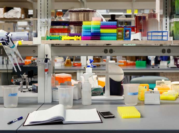 A lab notebook and supplies are shown on a lab bench. Boxes of supplies and lab equipment are shown in the background.
