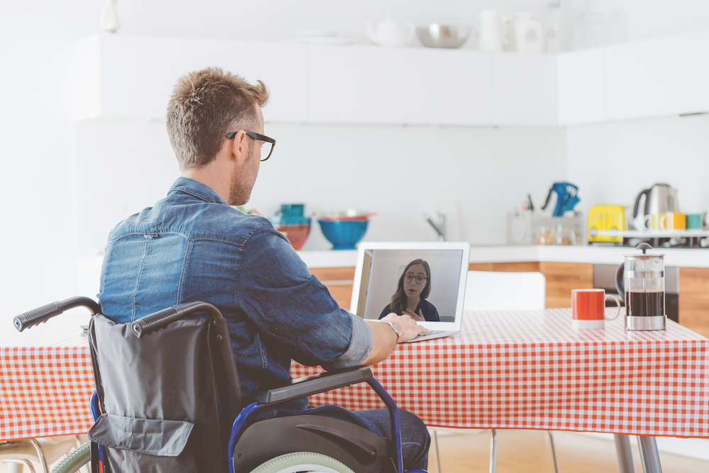 Back view of disabled man wearing jeans shirt sitting in a wheelchair at the kitchen table at home, using a laptop.