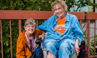 Two smiling women in their 80s; one is in an electric wheelchair, and the other is kneeling next to the wheelchair.