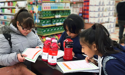 Three Hispanic tween girls stand around a selection of sugar-sweetened beverages with worksheets. One leans in to read the label on one of the drinks.