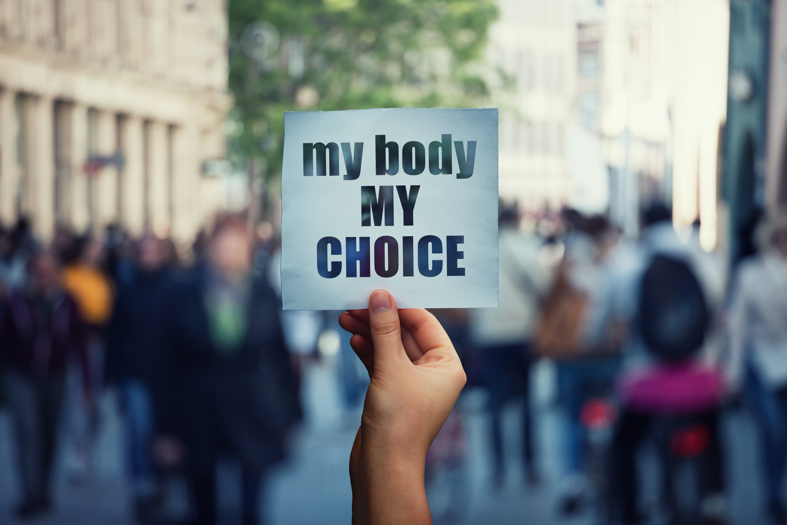 Feminist hands holding a protest banner with the message my body my choice over a crowded street.