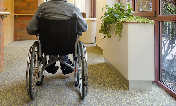 An elderly man, senior, is sitting in a wheelchair in the corridor near the window with his back to the camera. Nursing home.