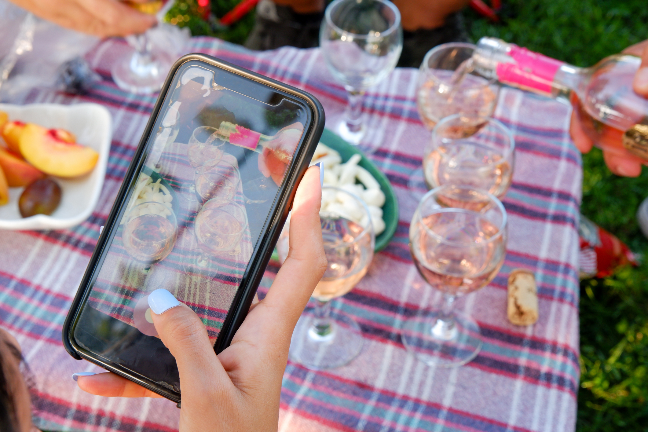 Woman's hand taking a photo of glasses of rose wine and cheese board with her smartphone on a picnic table