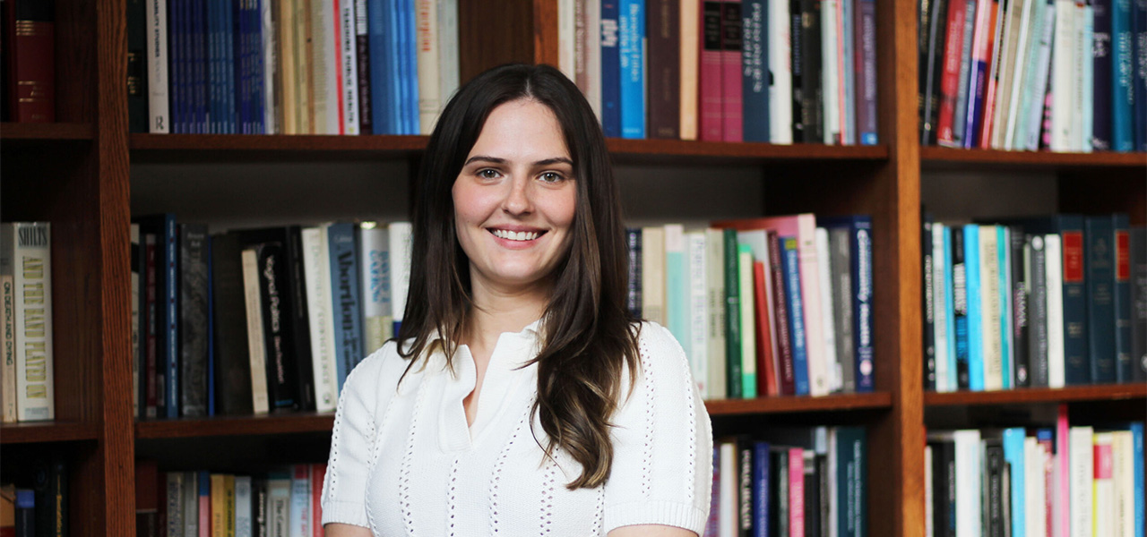 Dana Vasers, wearing a white, short-sleeved sweater, stands in front of a large bookcase and smiles at the camera