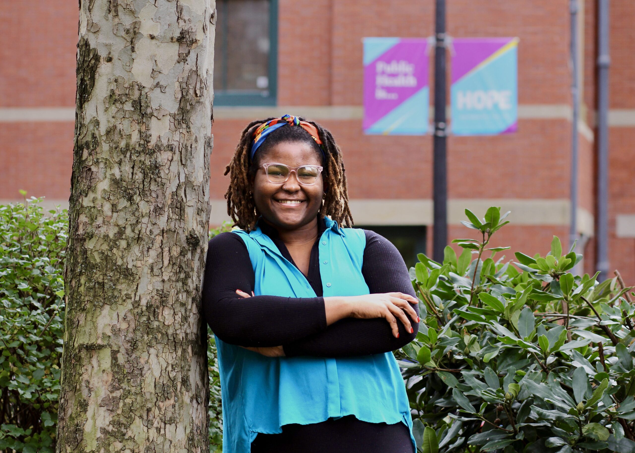 Almira Lewis, wearing a long black top and a bright blue blouse, leans against a tree on campus. A lamp post in the background says "Public Health is... HOPE."
