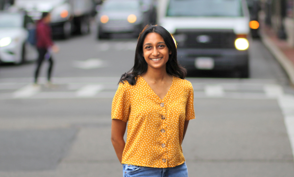 Nickita Gupta, wearing a mustard yellow blouse and blue jeans, stands at the crossroads of two streets in the South End