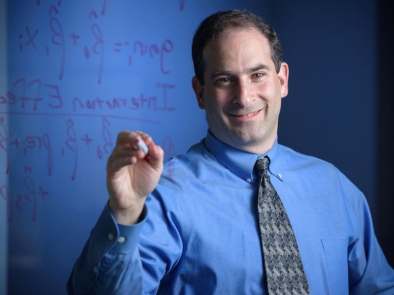 Headshot of alumnus Michael Posner holding a dry erase marker to a transparent board with statistical equations superimposed over his left shoulder