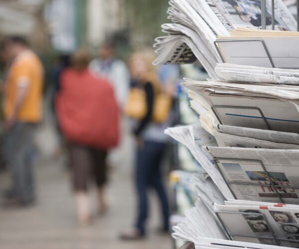 Newspaper rack on street, blurred people walk in the background