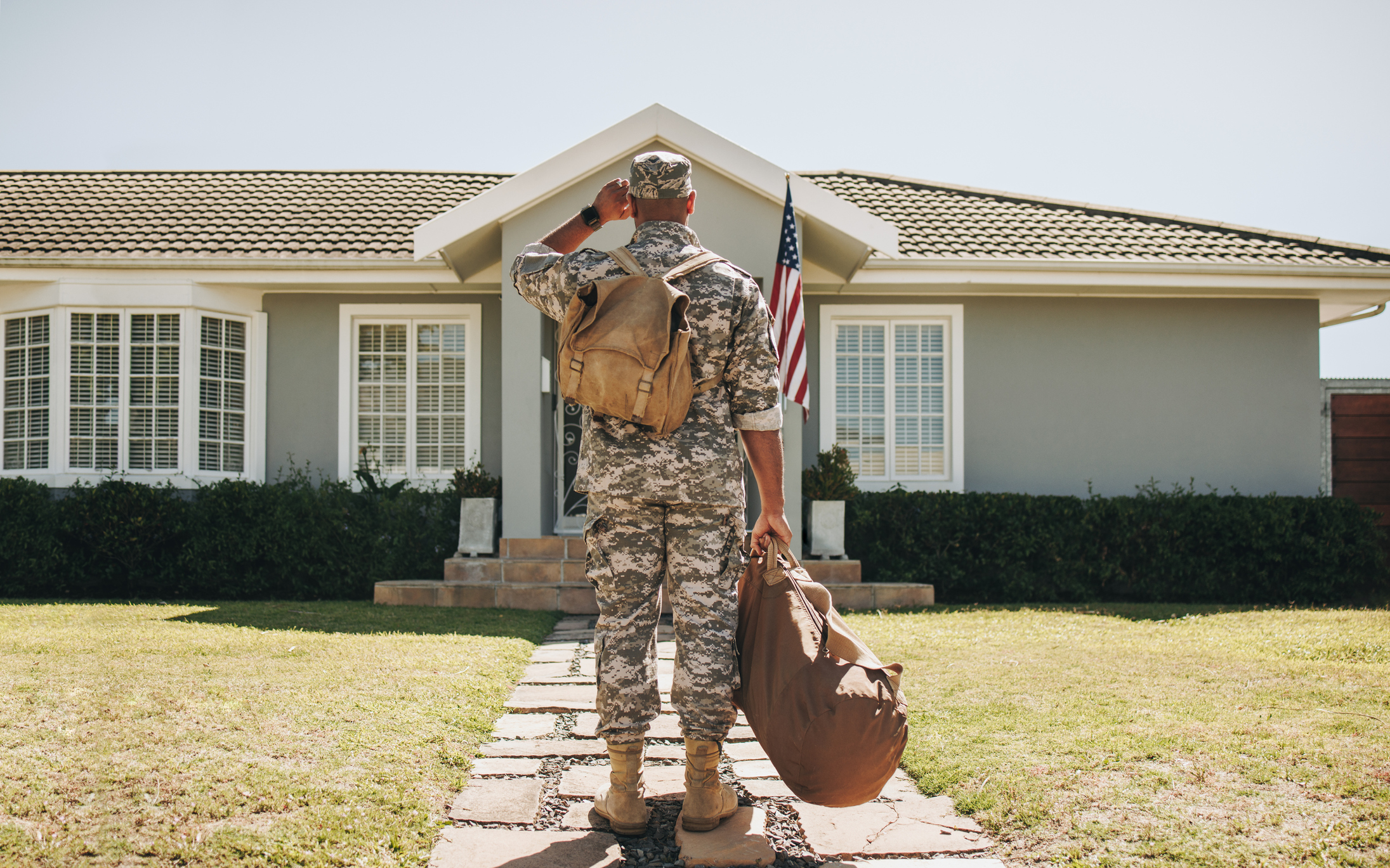 Rearview of a soldier returning home from the army. He is standing in front of his home with his luggage