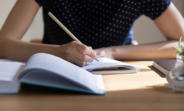 Young student girl preparing for college test, exam, writing notes, making draft, summary of textbook, studying, reading book for class report. Learning workplace table close up, banner shot