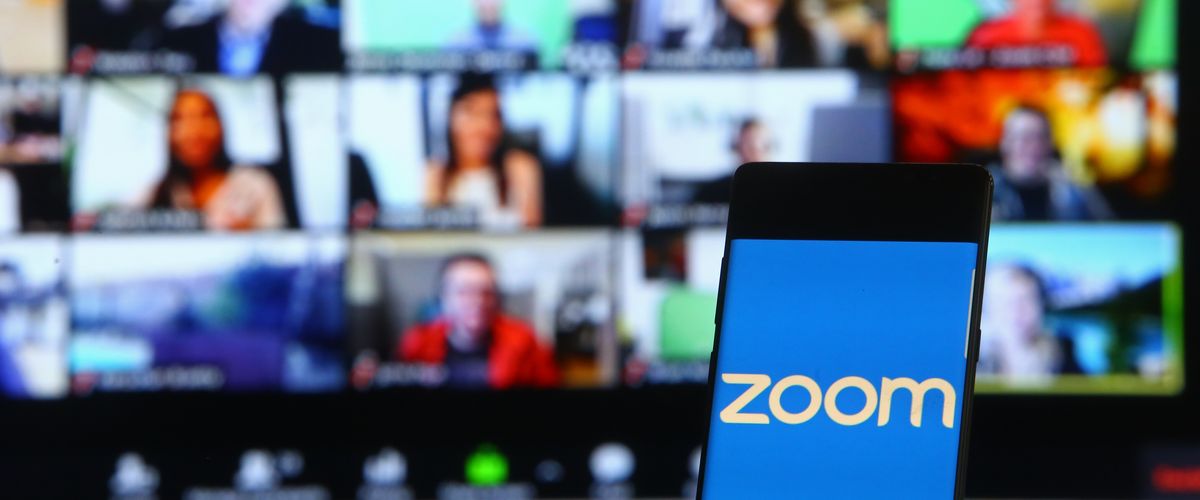 A phone showing the Zoom logo in front of a laptop screen showing many people in a video conference