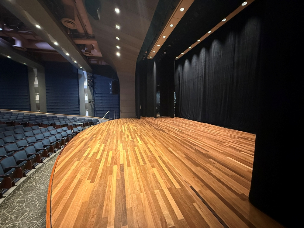 Side view of wood-floor stage with black curtains, with blue audience seats to the left.