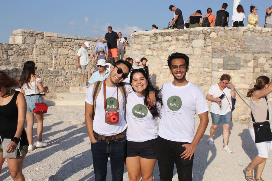 Shows three smiling young people standing in front of a stone wall