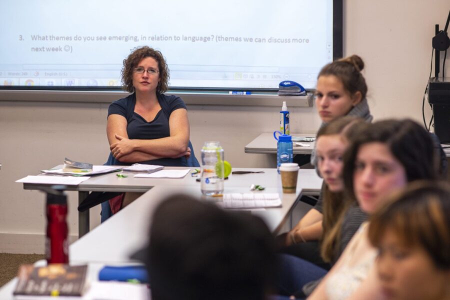 A photo of a professor sitting at a desk in front of her class.