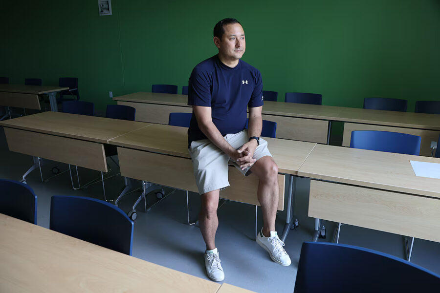 A photo of a man sitting on top of a desk in a classroom.