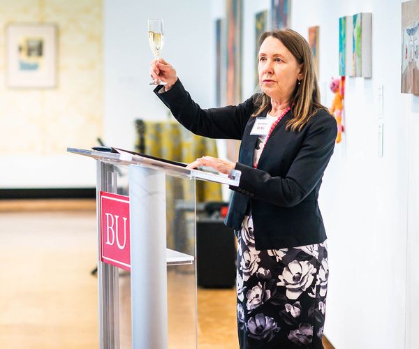 Photo: Dr. Beverly Brown, a white woman wearing a white blouse, black blazer, and black and white floral skirt, holds up a glass of champagne as she stands at a glass podium and addresses a crowd during a reception.